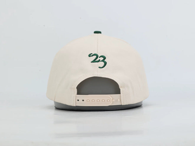 A cream-colored snapback cap shown from the back, featuring a green embroidered '23' on the back panel and a green button on top. The cap is displayed on a gray stand against a clean white background.