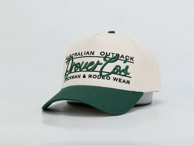 A baseball cap with an off-white crown and a dark green brim, featuring green and black embroidered text that reads 'Australian Outback Drovers' and 'Stockman & Rodeo Wear'.
