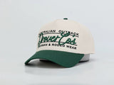 A baseball cap with an off-white crown and a dark green brim, featuring green and black embroidered text that reads 'Australian Outback Drovers' and 'Stockman & Rodeo Wear'.
