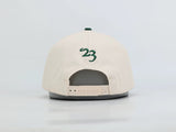 A cream-colored snapback cap shown from the back, featuring a green embroidered '23' on the back panel and a green button on top. The cap is displayed on a gray stand against a clean white background.